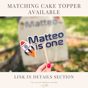 Pu&ograve; includere: Un topper per torta blu e bianco con un razzo e il testo "Matteo ha un anno".