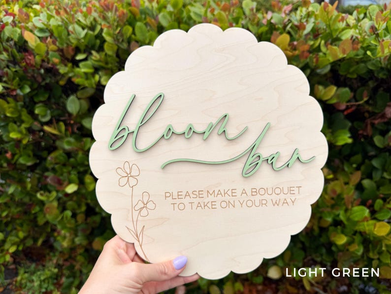 Pu&ograve; includere: Un cartello in legno verde chiaro con bordi a festoni che dice "Bloom Bar" in un font corsivo. Il cartello ha anche un piccolo disegno floreale e il testo "Per favore, crea un bouquet da portare via".