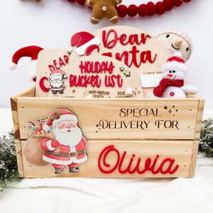 May include: A festive wooden crate with the words "Dear Santa" and "Special Delivery For Olivia" in red. Inside, a snowman, Santa Claus stickers, and a "Holiday Bucket List" sign. The crate is made of wood.