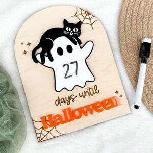 K&ouml;nnte beinhalten: H&ouml;lzernes Halloween-Countdown-Schild mit einer schwarzen Katze auf einem wei&szlig;en Geist, der die Zahl 27 anzeigt. Die W&ouml;rter 'Tage bis Halloween' und 'Halloween' sind orange. Enth&auml;lt einen schwarz-wei&szlig;en Marker.