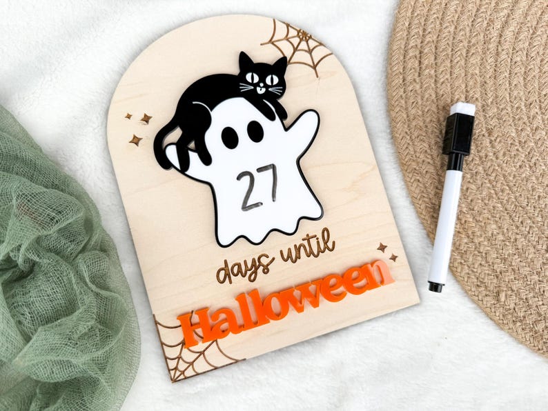 K&ouml;nnte beinhalten: H&ouml;lzernes Halloween-Countdown-Schild mit einer schwarzen Katze auf einem wei&szlig;en Geist, der die Zahl 27 anzeigt. Die W&ouml;rter 'Tage bis Halloween' und 'Halloween' sind orange. Enth&auml;lt einen schwarz-wei&szlig;en Marker.