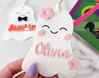 Op de afbeelding: Twee witte, spookvormige ornamenten met roze en oranje accenten. Het spook links heeft een zwarte strik en de naam "Austin" in oranje letters. Het spook rechts heeft roze bloemen en de naam "Olivia" in roze letters.