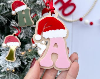 May include: Pink wooden letter A Christmas ornament with a red Santa hat on top. The ornament is hanging on a Christmas tree.