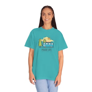 Puede incluir: Una camiseta azul turquesa con un diseño gráfico de una furgoneta azul con una tabla de surf en la parte superior, conduciendo por un bosque con un sol amarillo en el cielo. El texto "POUGE LIFE" está impreso en la camiseta, con las palabras "OUTDOOR GANG" debajo.