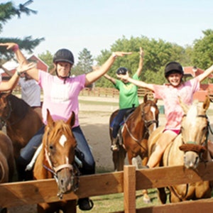 May include: A group of six people riding horses outdoors. They are all wearing helmets and smiling. The horses are brown and white. The people are wearing a variety of colourful clothing. The horses are in a fenced-in area.