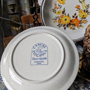 May include: A set of vintage stoneware plates with a floral design in yellow and orange. One plate is displayed with the underside facing up, revealing the "Casual Elegance Hearthside Stoneware Japan" stamp.