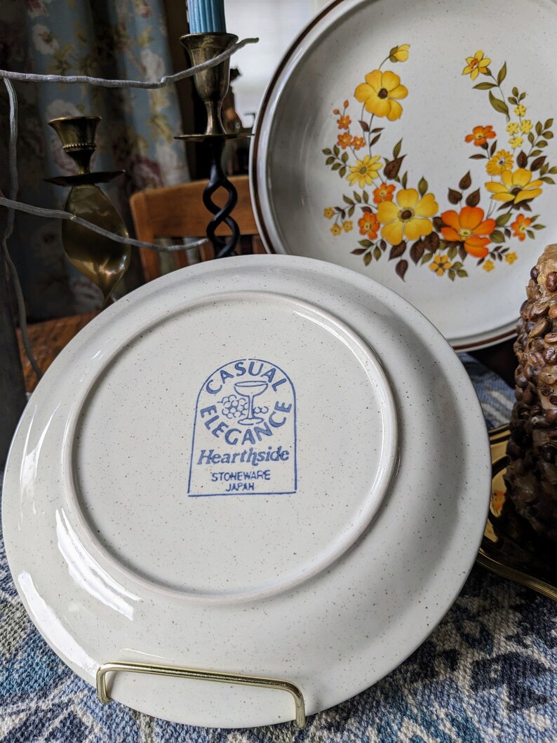 May include: A set of vintage stoneware plates with a floral design in yellow and orange. One plate is displayed with the underside facing up, revealing the "Casual Elegance Hearthside Stoneware Japan" stamp.