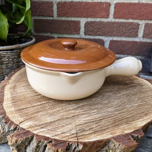 May include: A small, white ceramic casserole dish with a brown lid and a single handle. The dish is sitting on a wooden slice.