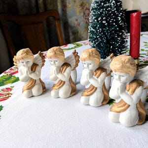 May include: Four ceramic angel figurines with folded hands in prayer. The angels have white bodies, gold accents on their wings and robes, and light brown hair. They are arranged on a white tablecloth with a Christmas-themed pattern.