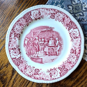 May include: A round white ceramic plate featuring a deep red transferware pattern. The rim is decorated with a dense floral motif, while the center depicts a historical scene of people gathered around a large bell suspended from a wooden frame.