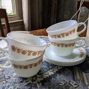 May include: Four white ceramic teacups with a brown floral pattern stacked on top of each other. The cups are on a blue and white patterned tablecloth.