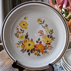 May include: A large, round ceramic plate with a floral design. The plate features a cream-colored speckled background with a brown rim and a vibrant arrangement of yellow and orange flowers. The plate is displayed on a wooden stand.