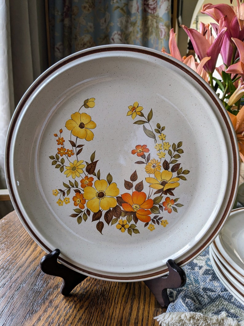 May include: A large, round ceramic plate with a floral design. The plate features a cream-colored speckled background with a brown rim and a vibrant arrangement of yellow and orange flowers. The plate is displayed on a wooden stand.
