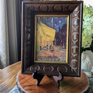 May include: A framed print of a painting depicting a cafe at night with a starry sky. The frame is dark brown wood with a carved design. The print is on a wooden easel.