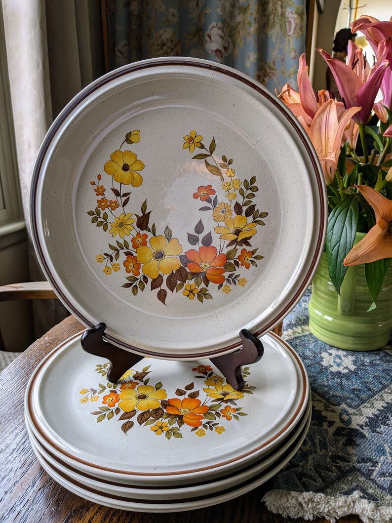 May include: A set of vintage ceramic plates with a floral design in yellow, orange, and brown. The plates have a speckled beige background and a brown rim. One plate is displayed on a wooden stand, with a stack of plates below.