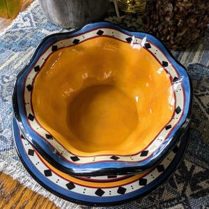 May include: A yellow ceramic bowl with a blue, black, and white rim. The bowl is sitting on a matching plate with a scalloped edge.
