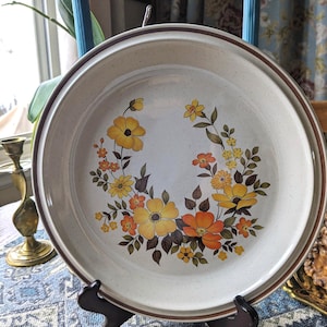 May include: A round, off-white ceramic plate with a brown rim and a floral design. The flowers are yellow and orange with brown leaves. The plate is displayed on a black stand.
