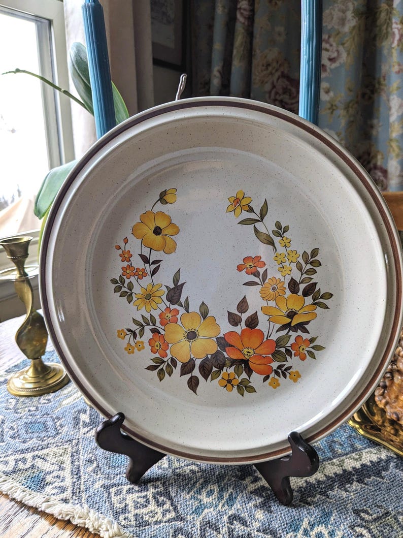 May include: A round, off-white ceramic plate with a brown rim and a floral design. The flowers are yellow and orange with brown leaves. The plate is displayed on a black stand.