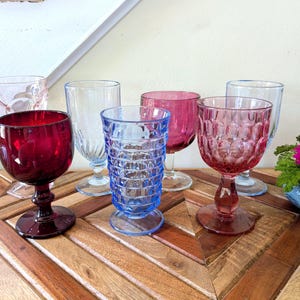 May include: A collection of colorful vintage wine glasses in shades of red, pink, and blue. The glasses have various textured designs and sit on a wooden surface. A vibrant pink flower in a blue bowl is also present.