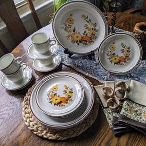 May include: A vintage dinnerware set featuring plates, bowls, cups, and saucers. The off-white pieces have a brown trim and a floral design with yellow and orange flowers. Napkins and napkin rings complete the set.