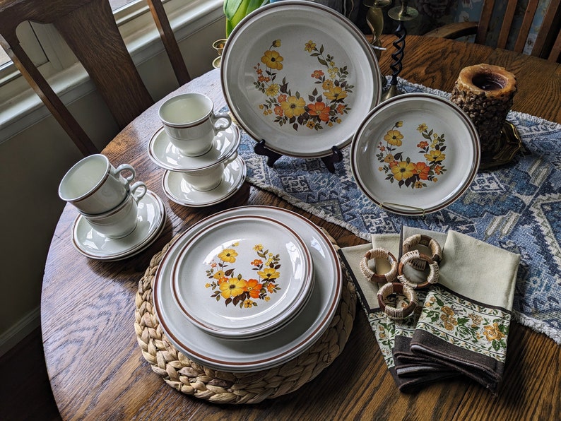 May include: A vintage dinnerware set featuring plates, bowls, cups, and saucers. The off-white pieces have a brown trim and a floral design with yellow and orange flowers. Napkins and napkin rings complete the set.