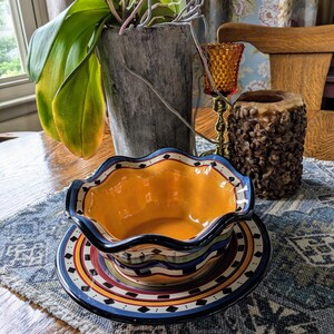 May include: A yellow and blue ceramic bowl with a wavy rim sits on a matching plate. The bowl and plate have a Southwestern-inspired design with black, red, and white accents. The bowl and plate are on a blue and white patterned placemat.