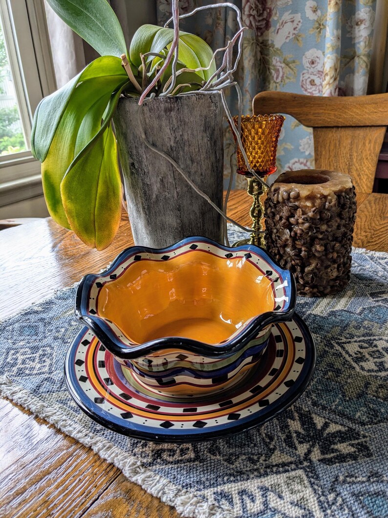 May include: A yellow and blue ceramic bowl with a wavy rim sits on a matching plate. The bowl and plate have a Southwestern-inspired design with black, red, and white accents. The bowl and plate are on a blue and white patterned placemat.