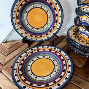 May include: Set of colorful ceramic dinner plates with a concentric circle design. The plates feature a yellow center with black, white, red, blue, and green rings.