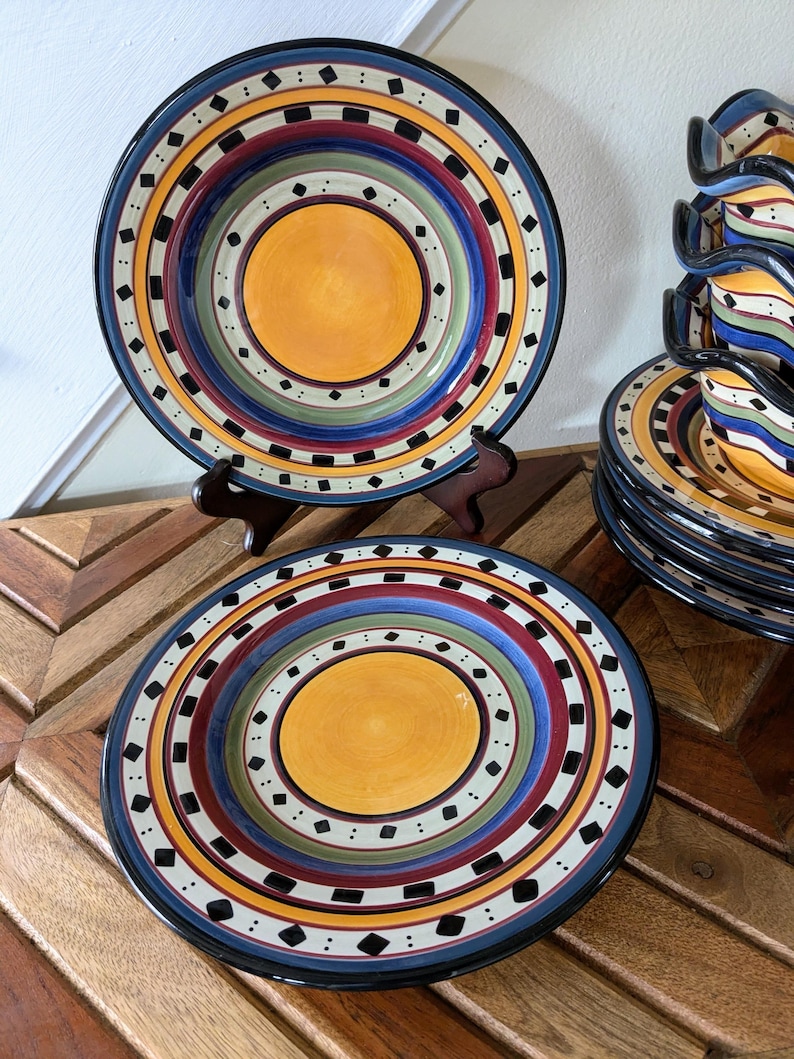 May include: Set of colorful ceramic dinner plates with a concentric circle design. The plates feature a yellow center with black, white, red, blue, and green rings.