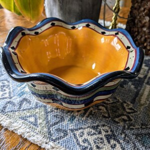May include: A yellow ceramic bowl with a scalloped rim and a blue, green, and black geometric design. The bowl is sitting on a blue and white patterned rug.
