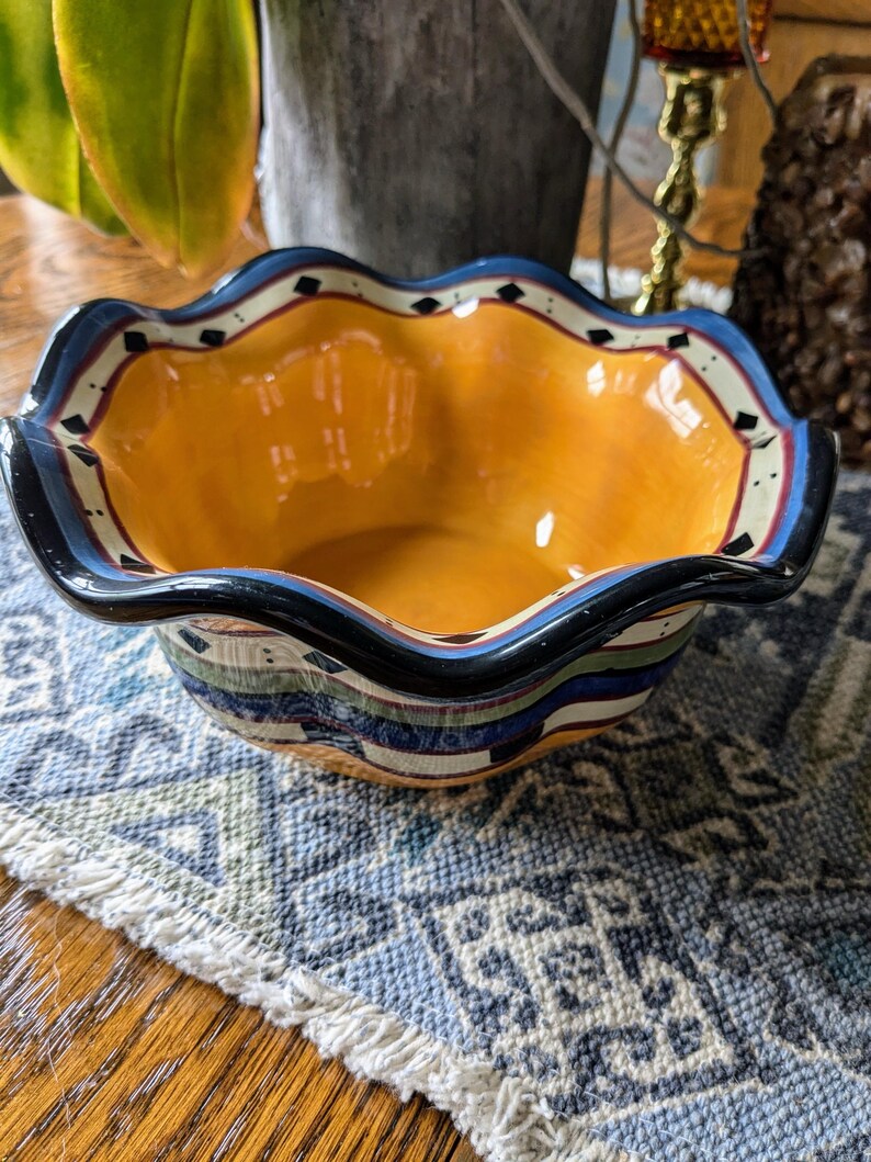 May include: A yellow ceramic bowl with a scalloped rim and a blue, green, and black geometric design. The bowl is sitting on a blue and white patterned rug.