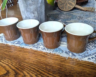 May include: Four brown ceramic mugs with white interiors are arranged on a blue and white patterned runner. The mugs have a classic design with a handle. The mugs are on a wooden table.