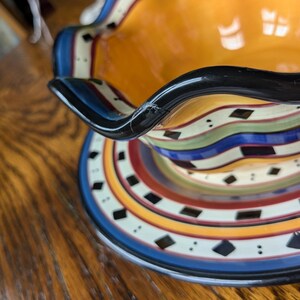 May include: A colorful ceramic serving bowl with a black rim and a wavy edge. The bowl has a multi-colored striped pattern with black diamond shapes.