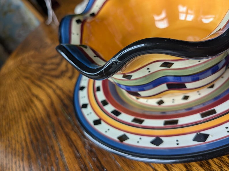 May include: A colorful ceramic serving bowl with a black rim and a wavy edge. The bowl has a multi-colored striped pattern with black diamond shapes.