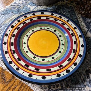 May include: A colorful ceramic dinner plate with a yellow center and concentric rings of red, blue, green, and white with black diamond shapes.