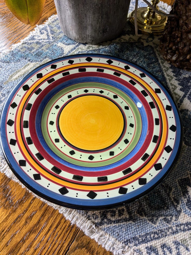 May include: A colorful ceramic dinner plate with a yellow center and concentric rings of red, blue, green, and white with black diamond shapes.