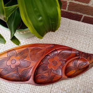 May include: A wooden serving tray with three sections, carved with floral designs and the word "HAWAII". The tray is brown and has a natural wood finish.