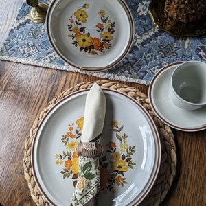 May include: A set of vintage ceramic dinnerware featuring a floral design in yellow and orange hues. The plates have a speckled, off-white background with a brown rim. A folded napkin with a patterned design sits on a plate.