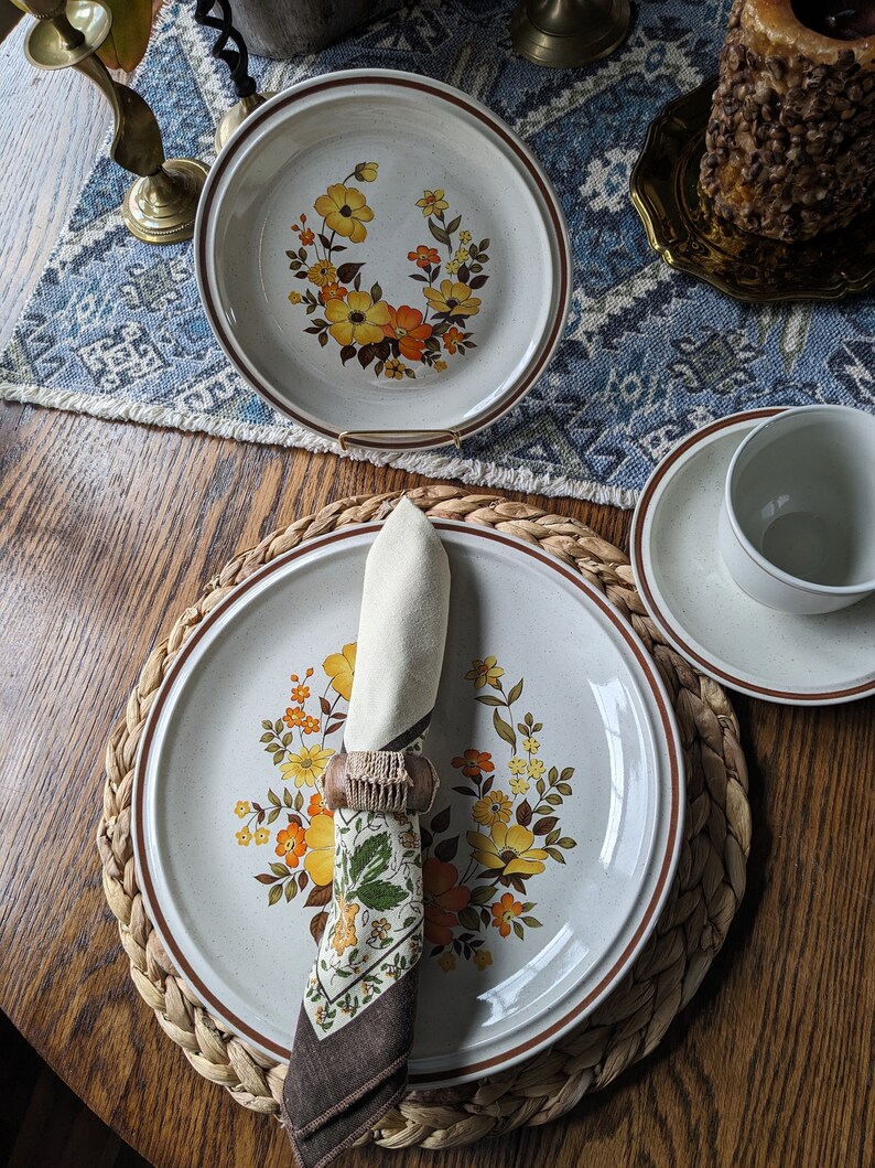 May include: A set of vintage ceramic dinnerware featuring a floral design in yellow and orange hues. The plates have a speckled, off-white background with a brown rim. A folded napkin with a patterned design sits on a plate.