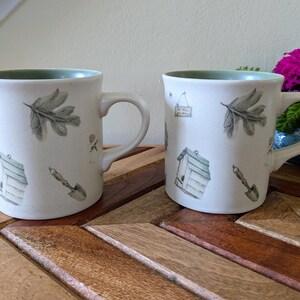 May include: Two ceramic mugs with a light beige exterior and a green interior. Each mug features illustrations of a leaf, a birdhouse, a trowel, and a small sign. The mugs are on a wooden table, with a vase of purple flowers in the background.