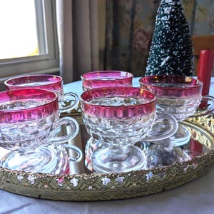 May include: A set of five clear glass teacups with a ruby red rim, resting on a decorative gold-colored tray. The cups have a diamond-cut pattern. A small Christmas tree and a red candle are in the background.