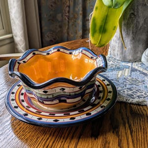 May include: A colorful ceramic bowl with a wavy rim and a matching saucer. The bowl is decorated with a blue, green, yellow, and red pattern. The saucer has a similar pattern with black diamonds.
