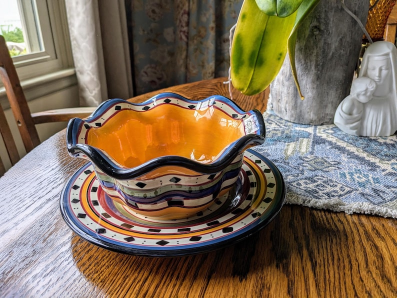 May include: A colorful ceramic bowl with a wavy rim and a matching saucer. The bowl is decorated with a blue, green, yellow, and red pattern. The saucer has a similar pattern with black diamonds.