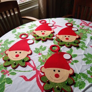 May include: Four felt Christmas elf ornaments with red hats, white faces, and green glitter collars. Each ornament has a white circle for writing a name.