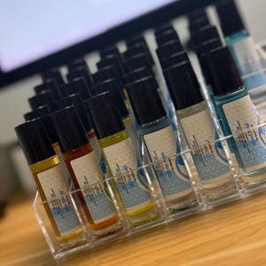 May include: A clear acrylic organizer holds several glass roll-on bottles with black caps. The bottles contain various colored liquids, including yellow, orange, and blue. Each bottle has a white label with blue text and a decorative pattern.