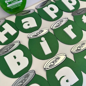 May include: Green and white paper cutouts of the word "Happy Birthday" in the shape of aluminum cans. The cans have a silver top and a green bottom. The word "BuzzBall" is visible on one of the cans.