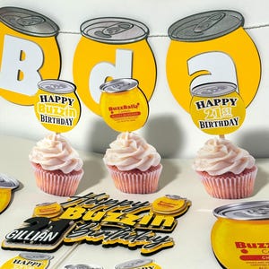 May include: A 21st birthday party decoration set with a yellow and black color scheme. The set includes a cake topper, cupcake toppers, and a banner with the words "Happy Buzzin Birthday".