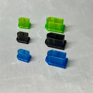 May include: An assortment of small, rectangular, 3D-printed objects in green, black, and blue. The objects have a box-like shape with a base. The translucent green and blue pieces are next to solid black pieces.
