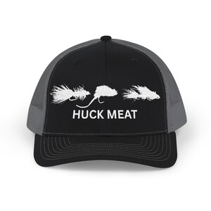 Può includere: Cappellino da camionista nero e grigio con visiera curva. Il cappellino presenta la scritta bianca "HUCK MEAT" e tre esche da pesca a mosca bianche. Il retro è in rete per la ventilazione.
