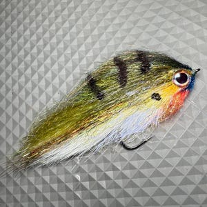 May include: A colorful fishing lure designed to resemble a fish. The lure features a combination of green, yellow, and white fibers, with black stripes and a red, yellow, and blue head. A black hook is attached.